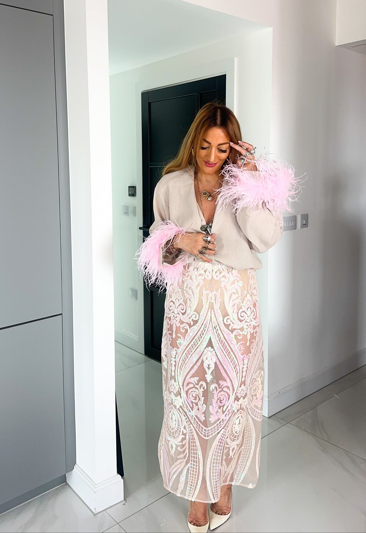 A woman wearing a beige cardigan with pink feathers and a matching beige and pink embroidered midi skirt, standing in a modern white hallway.