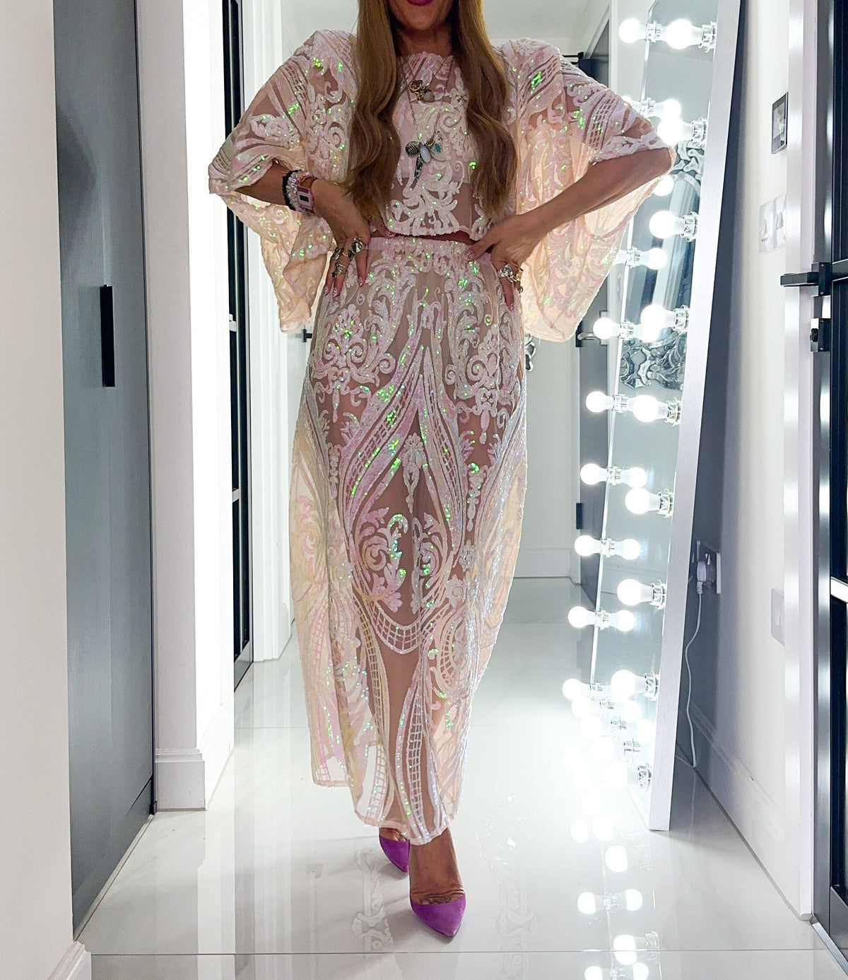A woman posing in a hallway wearing a long, sheer beige maxi dress with intricate green and white paisley embroidery, paired with purple heels.