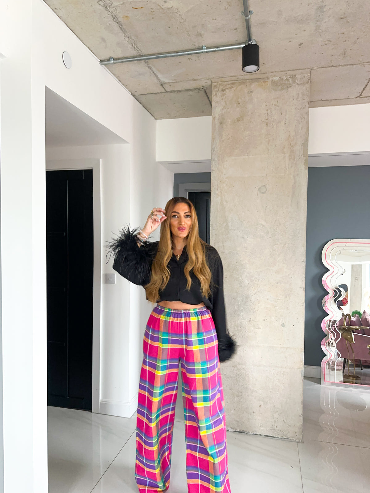 A woman stands in a modern indoor space wearing a black cropped long-sleeve top with feathered cuffs and bright pink plaid wide-leg pants with yellow, blue, and green lines.