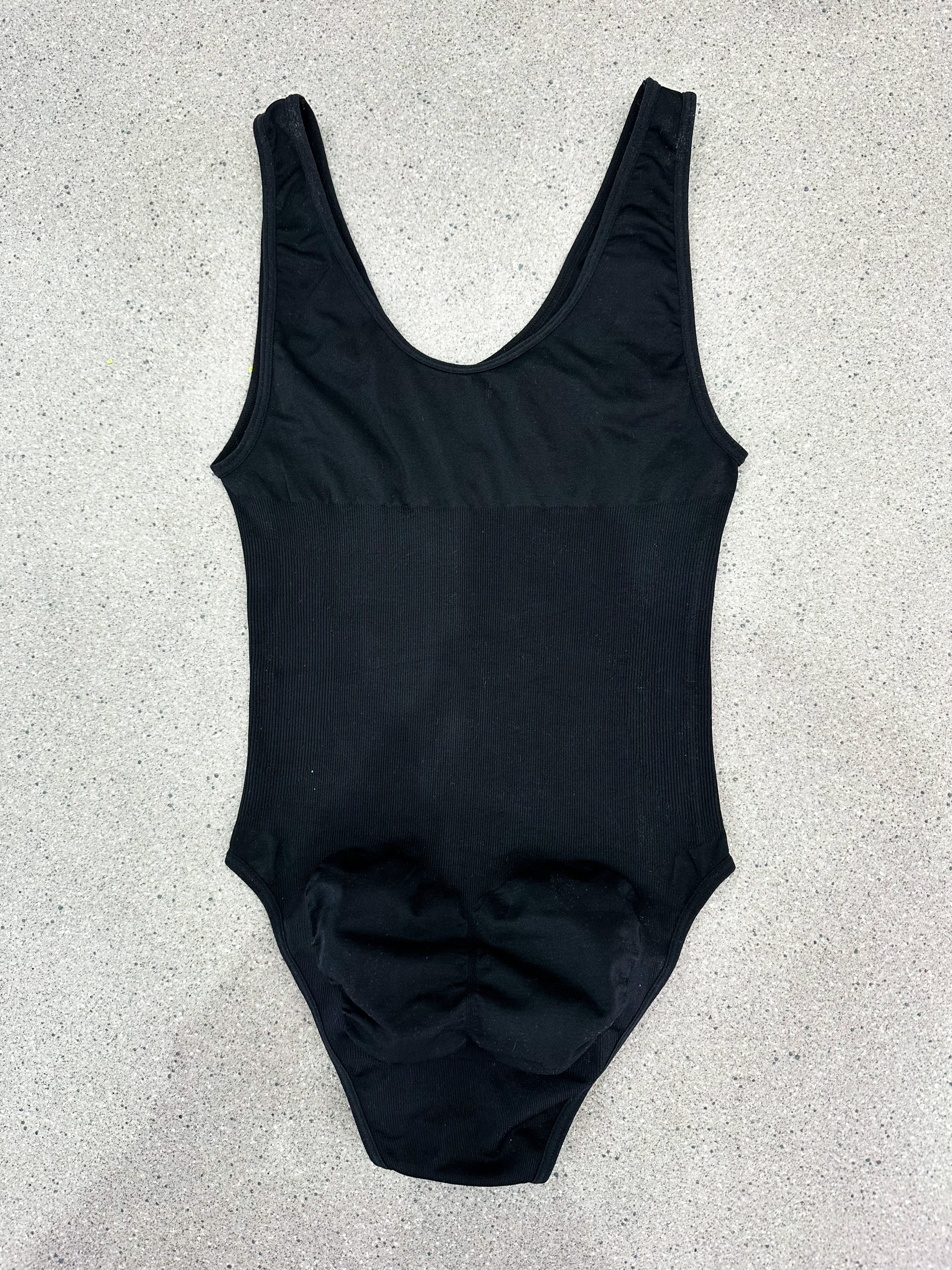 A black sleeveless bodysuit or one-piece swimsuit laid flat on a light gray speckled surface.