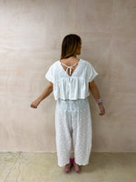 back of model wearing Embroidered Pleated Hem Swing style cropped Top In white with t-shirt style sleeves styled with Broderie Anglaise lace trousers in white