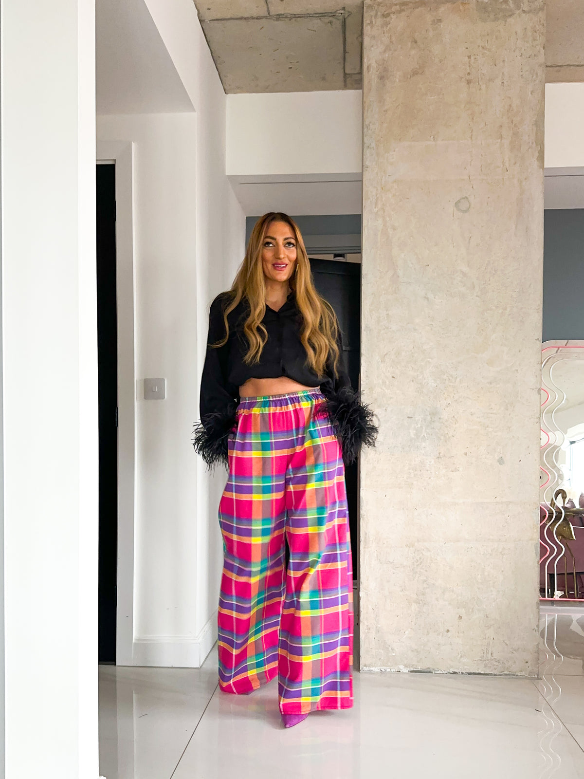 A woman stands in a modern indoor hallway wearing a black cropped long-sleeve top with black feather trim and bright pink, yellow, and blue plaid wide-leg pants.