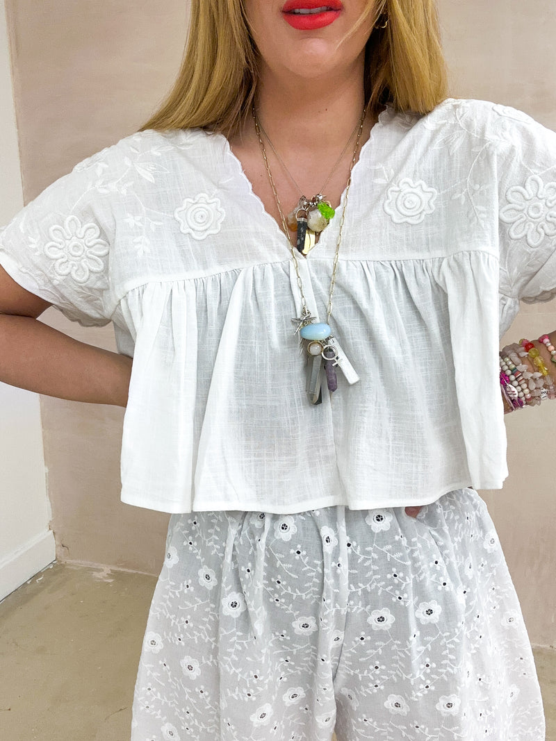 close up of model wearing Embroidered Pleated Hem Swing style cropped Top In white with t-shirt style sleeves styled with Broderie Anglaise lace trousers in white