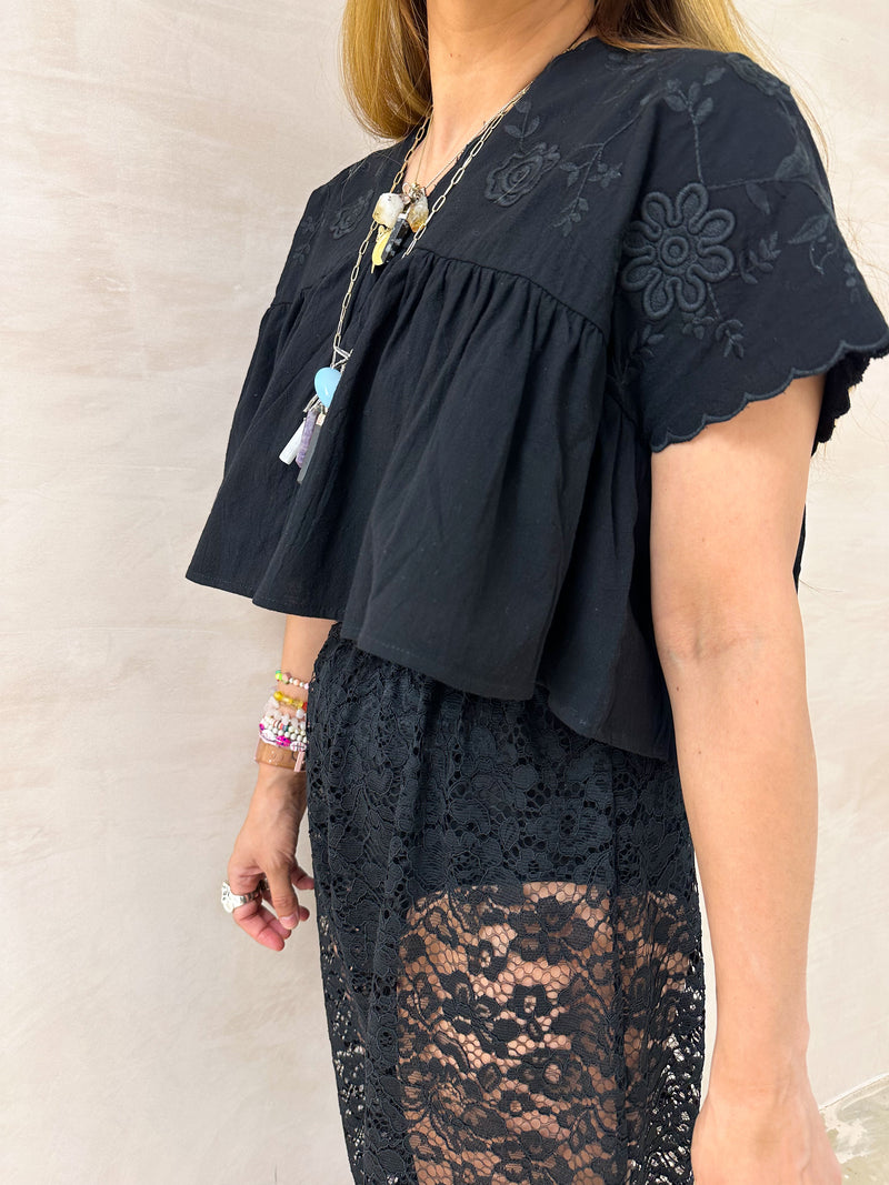 close up of model wearing Embroidered Pleated Hem Swing style cropped Top In Black with t-shirt style sleeves styled with ballon lace trousers in black 