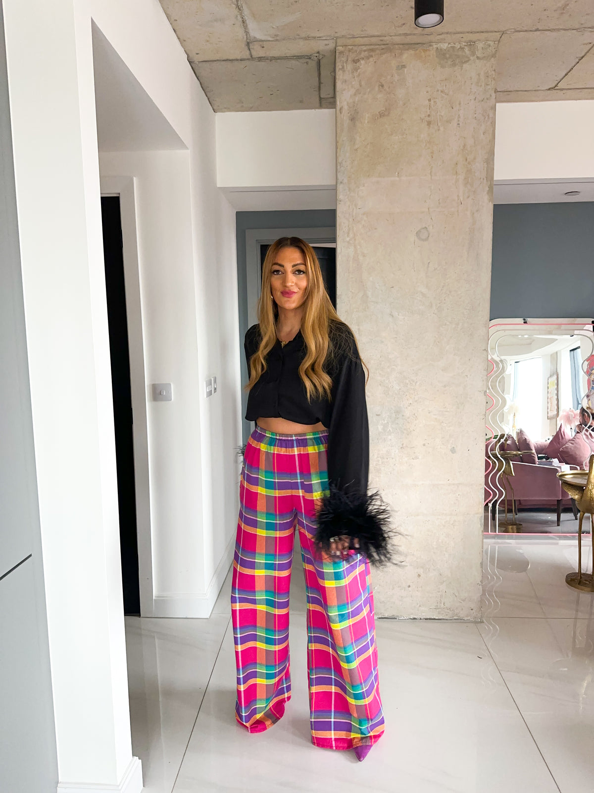 A woman stands in a modern indoor hallway wearing bright multicolor plaid wide-leg pants with pink, yellow, blue, and green tones, paired with a black cropped long-sleeve top and holding a black feather-trimmed clutch.