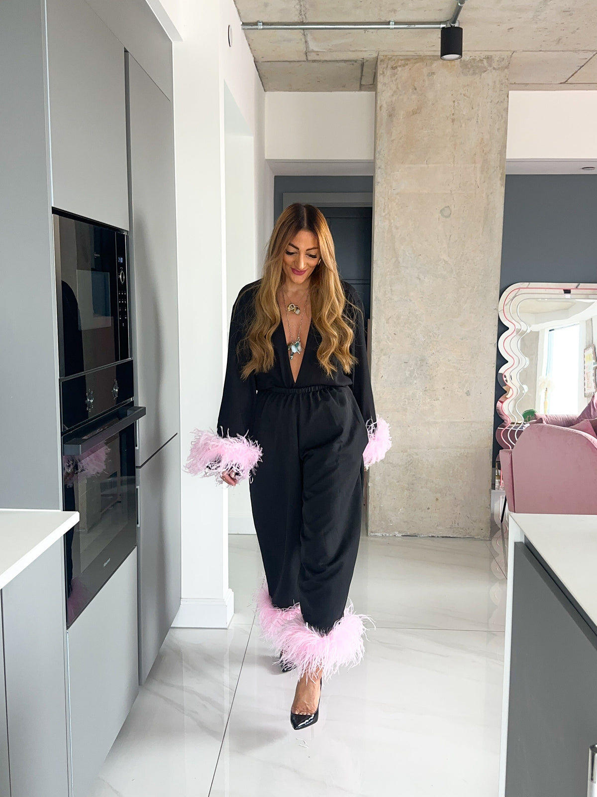A woman wearing a black jumpsuit with fluffy light pink feather trim on the cuffs and hem, walking in a modern kitchen hallway.